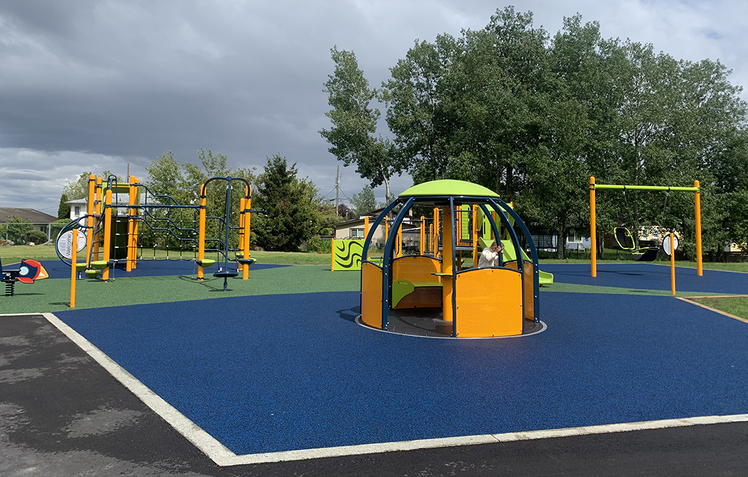 Photo of a playground with a rubberized surface and orange, green, and blue playground equipment, including a We-Go-Round merry-go-ground in the foreground, and a friendship swing and play structure in the background. There is a green leafy tree and a grey sky in the background.