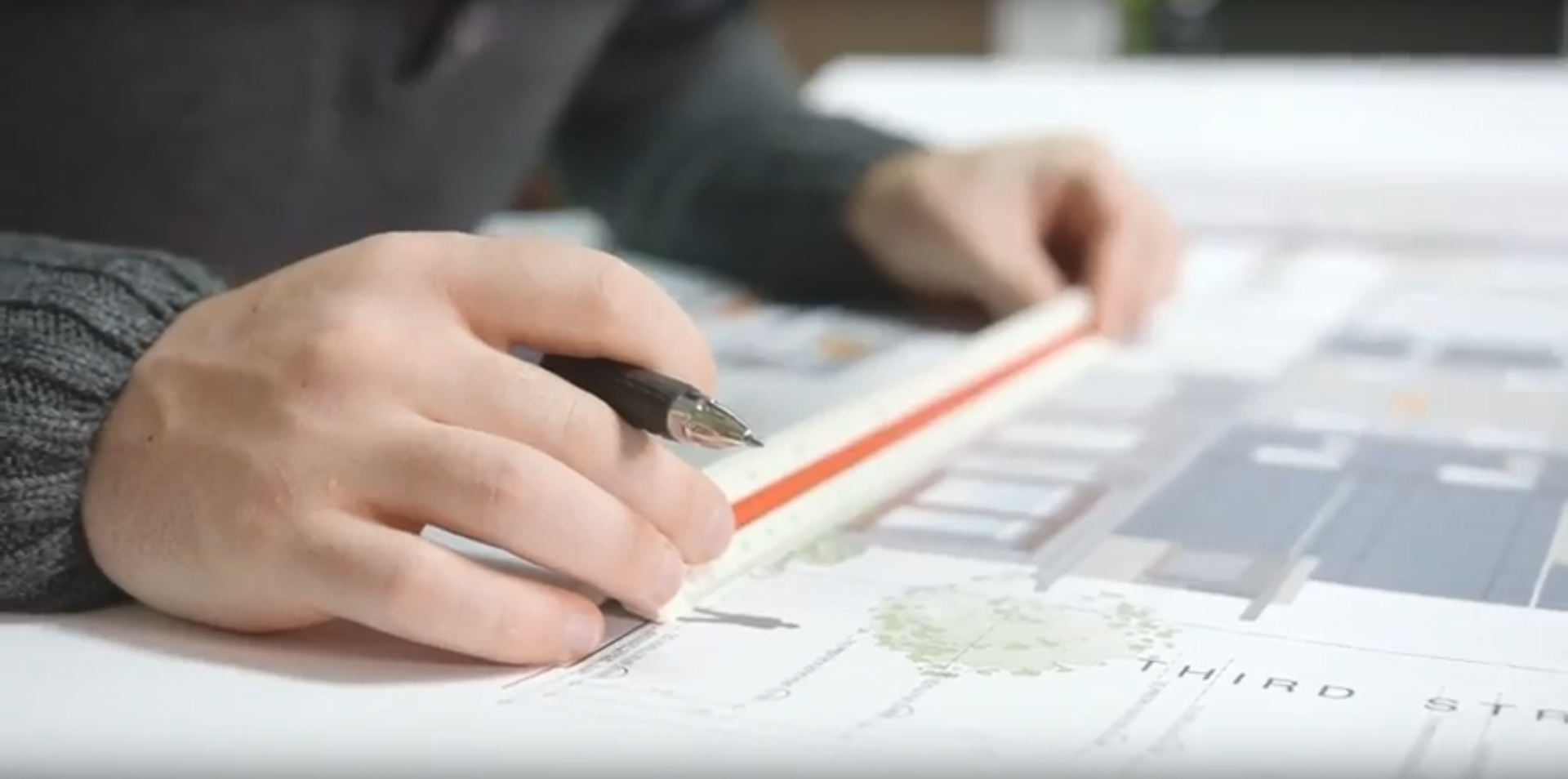 Photo of a municipal planner reviewing building plans. The planner's hands are holding a pen and a ruler and are resting on a set of plans depicting a residential building.