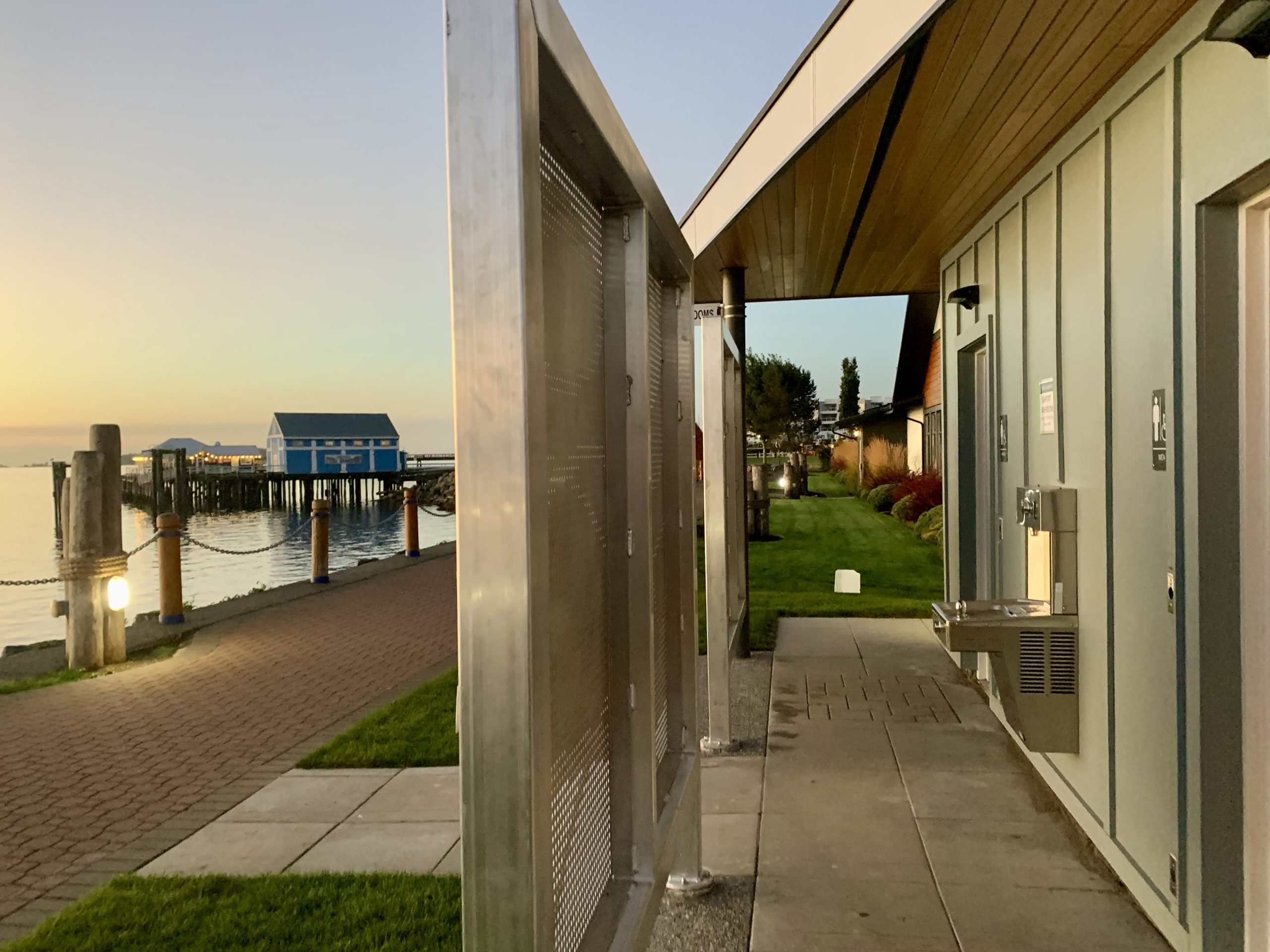 Side view of the Seaport Washrooms, with the accessible men's washroom door and accessible women's washroom door visible, and a water fountain with a water bottle refilling station between the two doors. A metal privacy screen separates the washroom doors from the bricklock waterfront walkway, and the Salish Sea, Beacon Wharf, and Fishmarket are in the background.