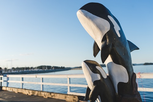 A wood sculpture of a mother and baby orca along the waterfront at sunset.