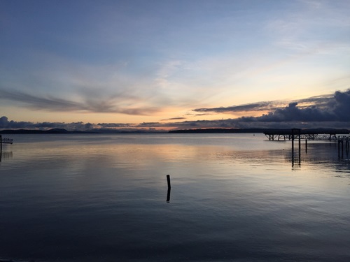 A sunrise over a calm waterfront. The fishing pier in the background reflects clearly in the water, as does a lone log.