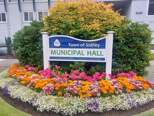 A large wooden sign with the top shaped lake a wave, reading "Town of Sidney Municipal Hall". It sits in a vibrant flower bed full of pink, white, orange, and purple flowers, with a backdrop of leafy green bushes.