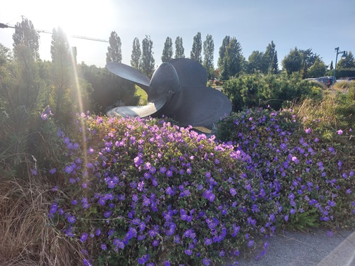 An old ship propeller sits on display in the middle of a purple and green flower bed.