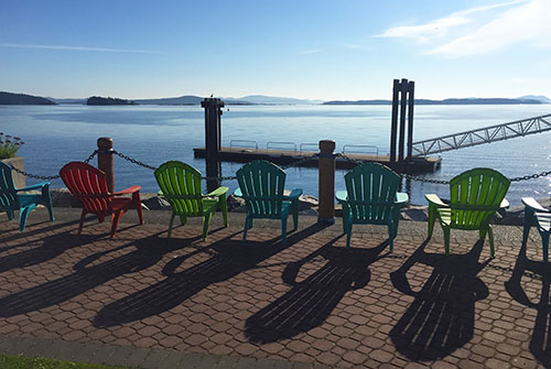 Plastic Adirondack chairs face the Salish Sea, bird perches, and a barge, along the waterfront walkway. The chairs cast long shadows and are each their own colour: blue, red, green, blue, blue, green, and blue.