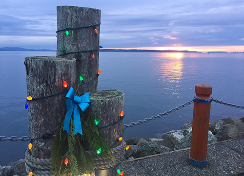 Sunrise over the Salish Sea, with a festively decorated bollard, complete with twinkle lights and a vibrant blue bow.